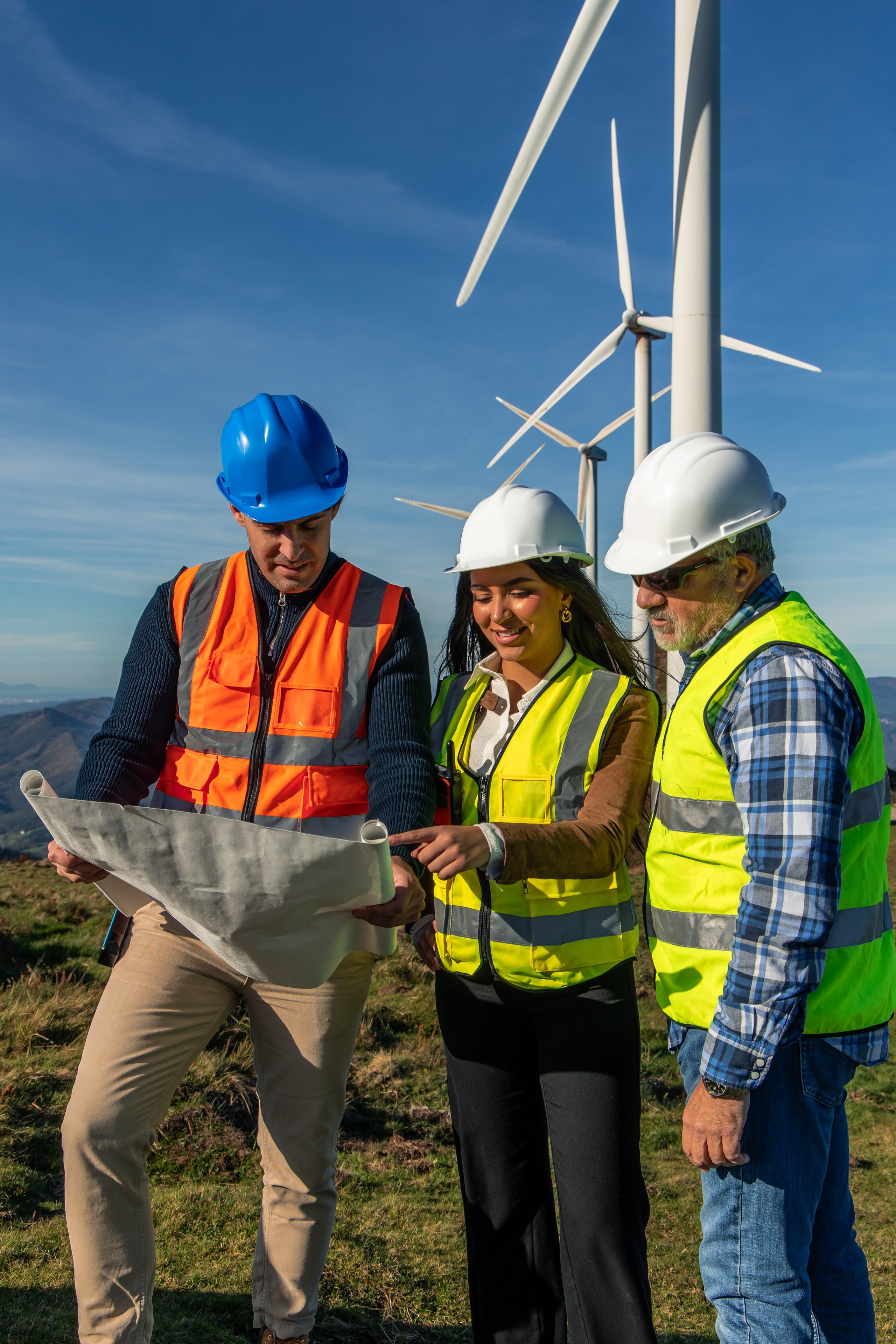 Three multiethnic engineers wearing safety vests and hard hats are examining a blueprint in front of a row of wind turbines